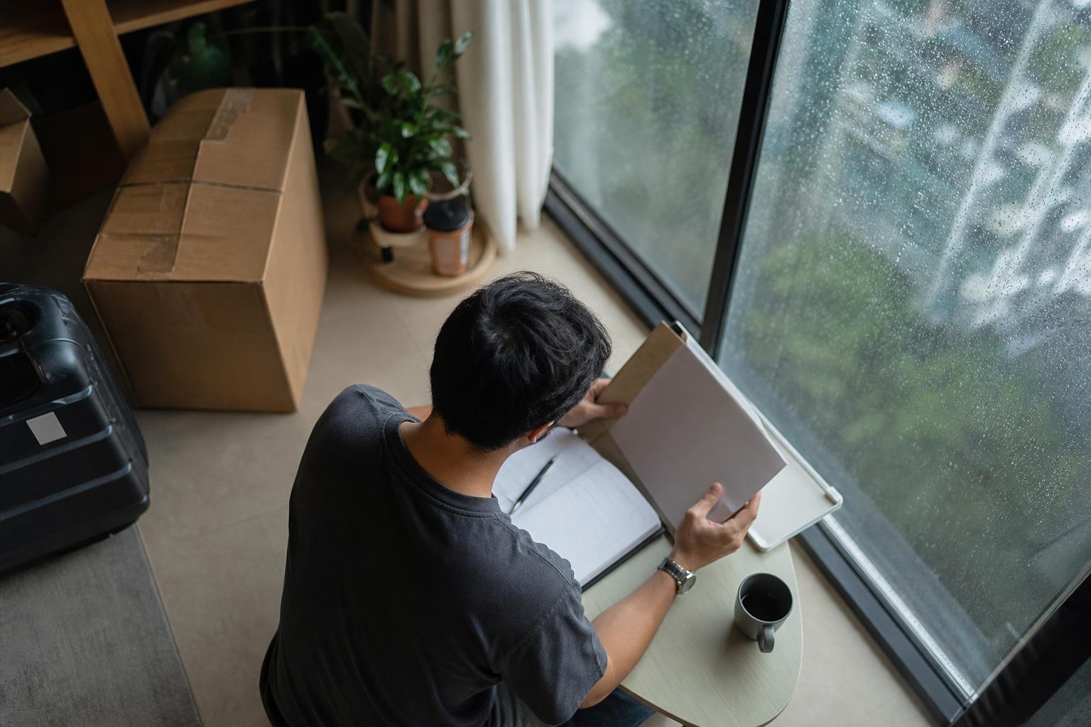 一名搬家中的男子在雨天窗边翻阅文件盒，桌上放着纸质记事本和笔