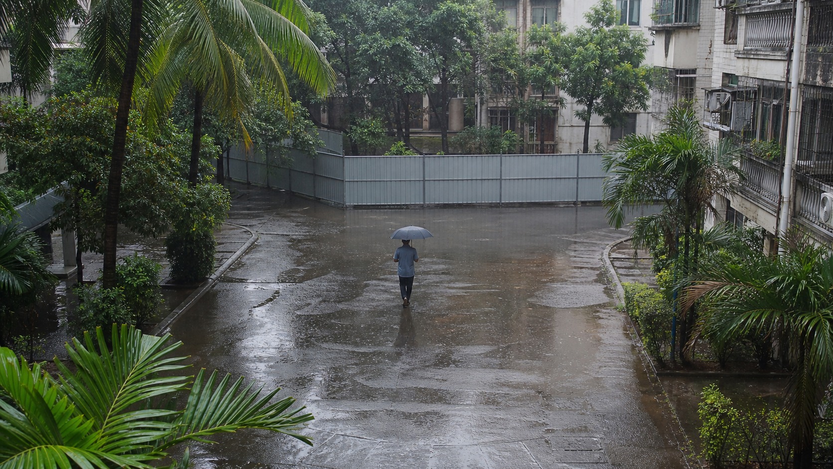 雨天的海口老旧住宅庭院里，一名住户撑伞走向施工围挡